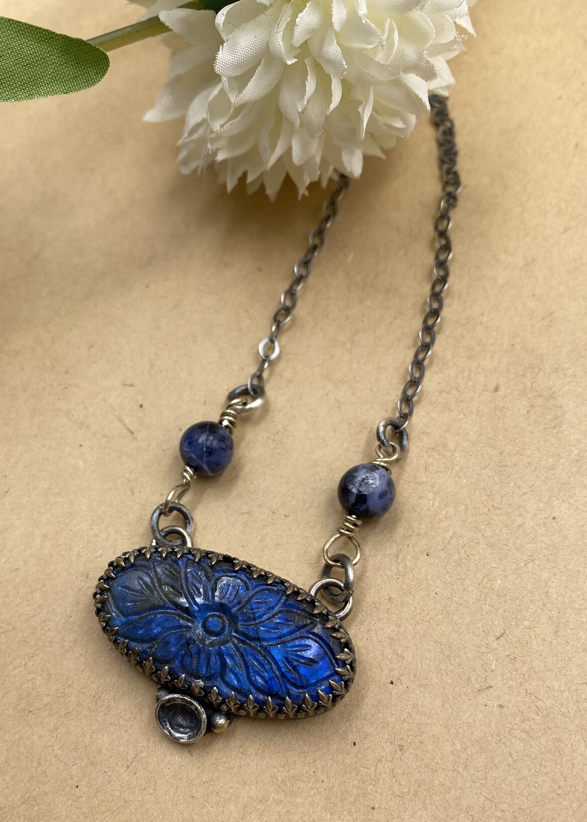 Oval Labradorite stone with flower carved in it below a Sterling silver chain with two blue stones partway up. Below the focal piece is a Sterling silver watercasting flanked by two small gold balls.