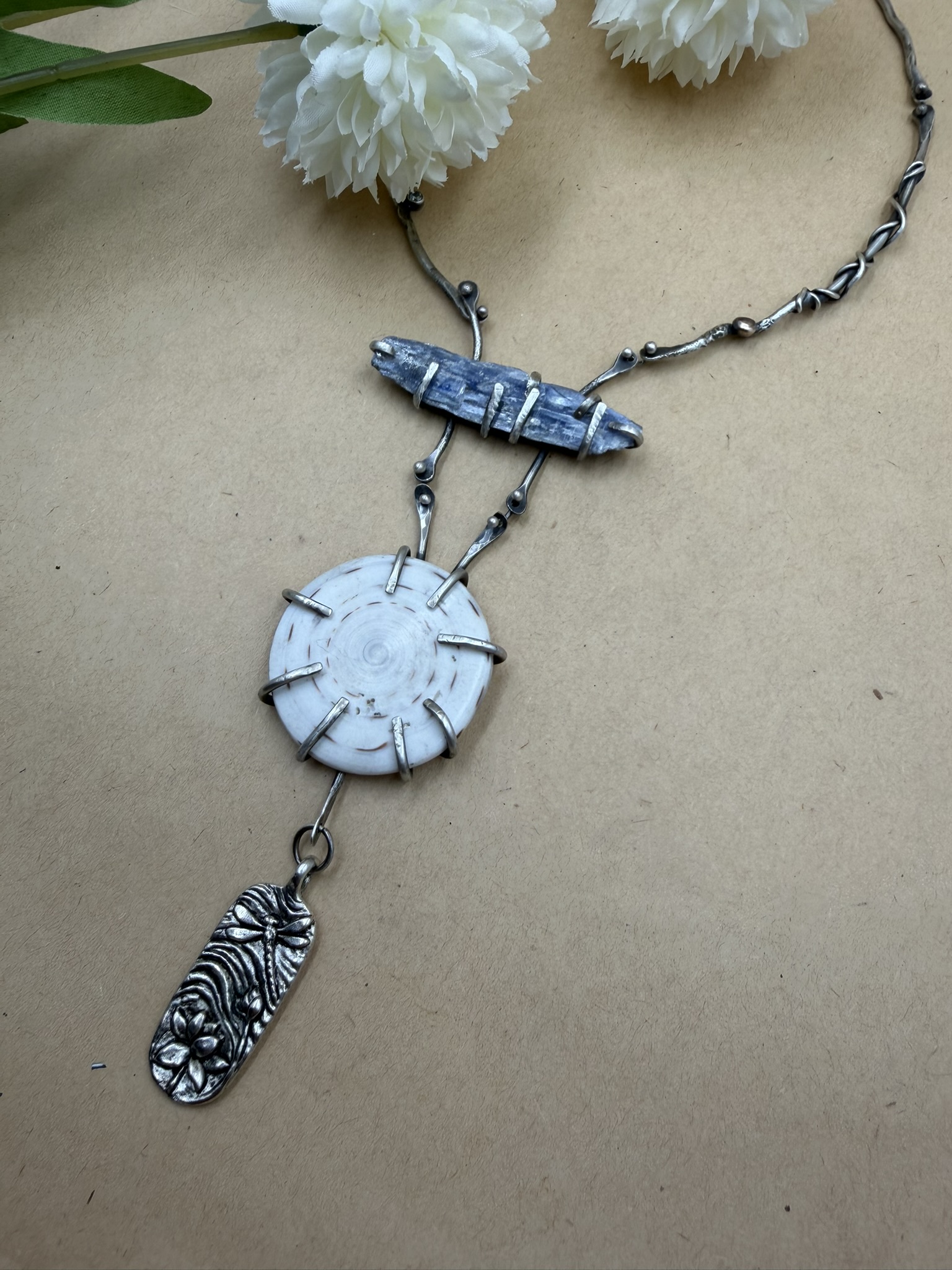 Sterling silver necklace featuring a horizontal piece of raw kyanite above a Shiva shell and a pewter dragonfly pendant. The necklace is of decorated silver wire pieces.